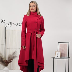 May include: A vibrant red coat featuring a high collar, three large circular buttons, and a waist-cinching tie belt. The garment has a high-low hemline that is shorter in the front and longer in the back, creating a distinct flared silhouette.