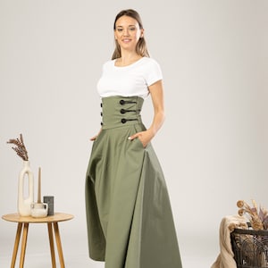 Może przedstawiać: Oliwkowa sp&oacute;dnica maxi z gorsetem z wysokim stanem i guzikami. Sp&oacute;dnica ma pełną, zwiewną sylwetkę i kieszenie. Modelka ma na sobie biały t-shirt. Stylowy dodatek do każdej garderoby.