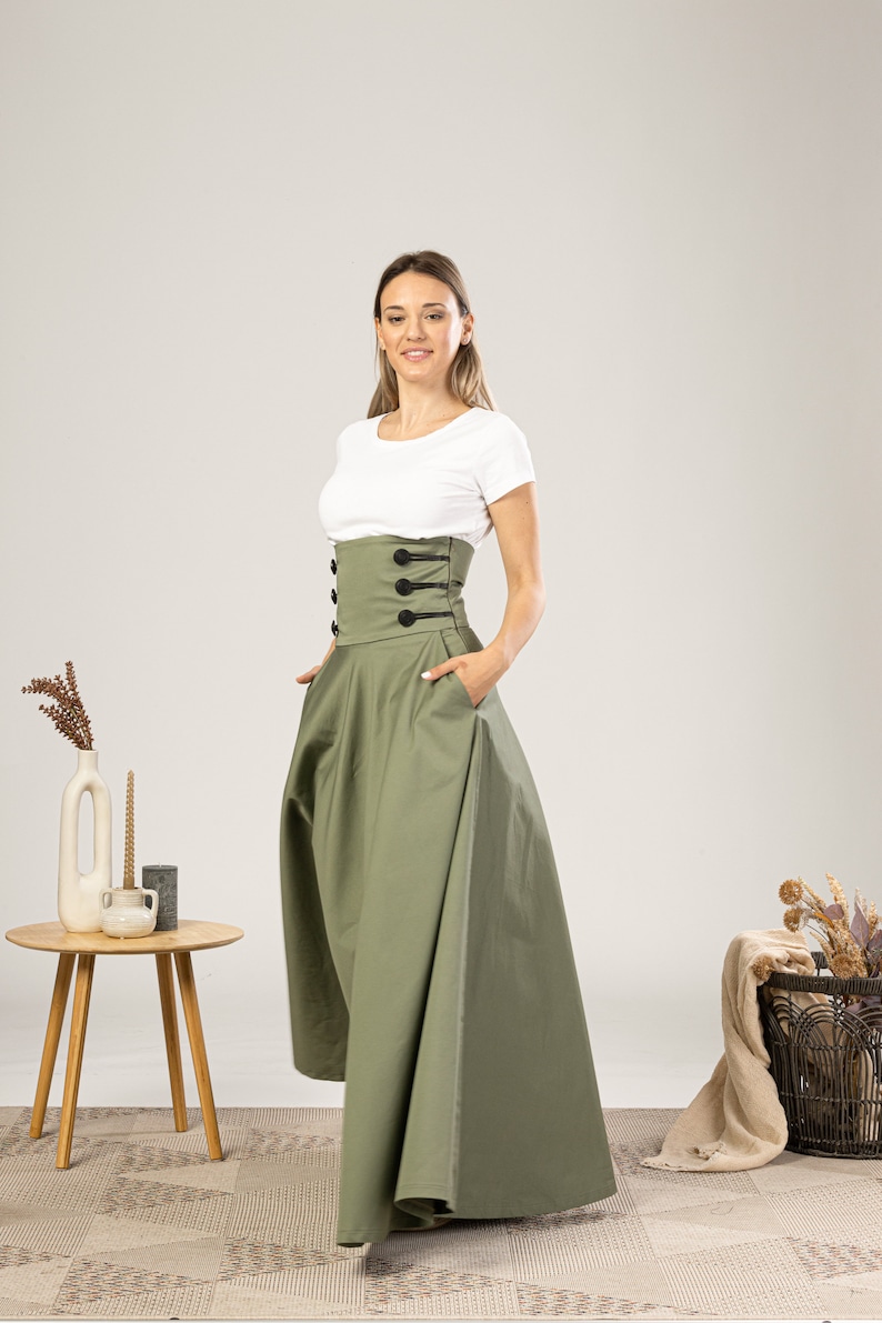 Może przedstawiać: Oliwkowa sp&oacute;dnica maxi z gorsetem z wysokim stanem i guzikami. Sp&oacute;dnica ma pełną, zwiewną sylwetkę i kieszenie. Modelka ma na sobie biały t-shirt. Stylowy dodatek do każdej garderoby.