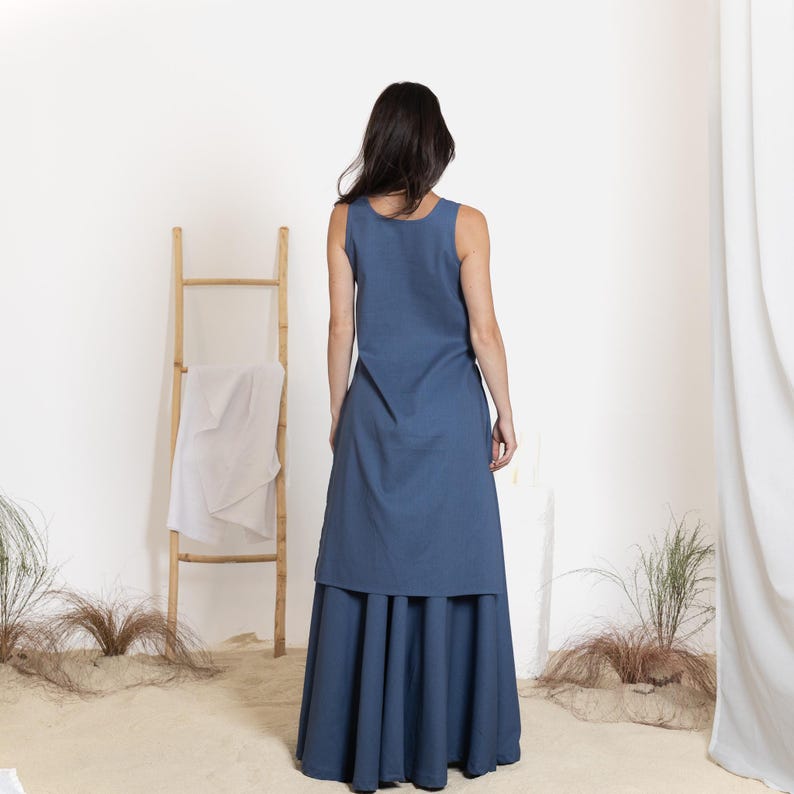 May include: A sleeveless, blue dress with a layered design. The top layer is a simple, straight cut, while the bottom layer is a pleated skirt. A wooden ladder and white cloth are in the background.