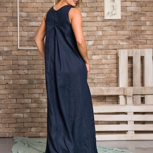 K&ouml;nnte beinhalten: Ein &auml;rmelloses, marineblaues Maxikleid mit einem plissierten R&uuml;ckendetail. Das Kleid hat ein schlichtes, elegantes Design und reicht bis zum Boden. Das Model steht vor einer Backsteinmauer und einem hellgr&uuml;nen Stoff.