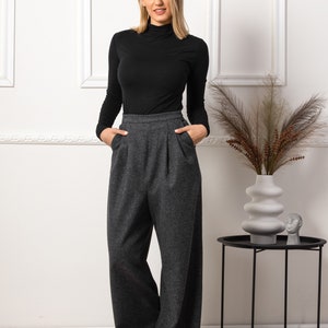 May include: A woman wearing a black turtleneck top and gray wide-leg trousers. The pants have a high waist and pleats. The outfit is paired with a decorative vase and a small side table.