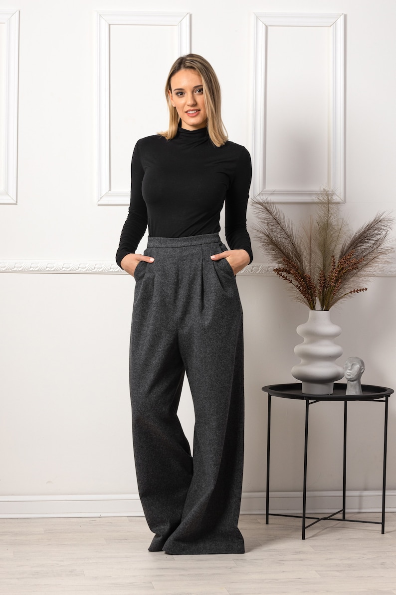 May include: A woman wearing a black turtleneck top and gray wide-leg trousers. The pants have a high waist and pleats. The outfit is paired with a decorative vase and a small side table.