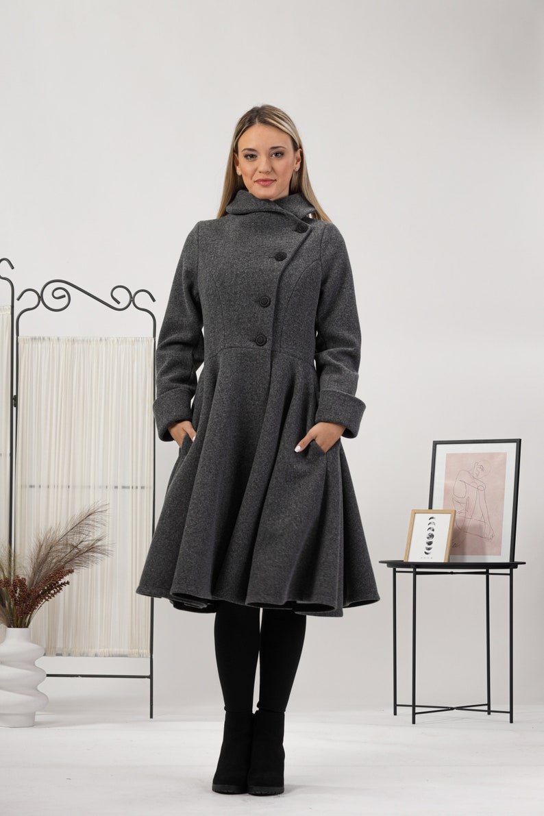 May include: A charcoal grey coat featuring a high collar, long sleeves with folded cuffs, and a flared, knee-length skirt. The coat has a button-down front closure with three visible black buttons and two side pockets integrated into the design.