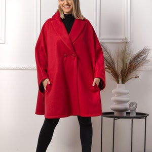 May include: A double-breasted red coat with large lapels and pockets. The coat is knee-length and worn with black leggings and ankle boots. The background is a white wall with decorative trim.