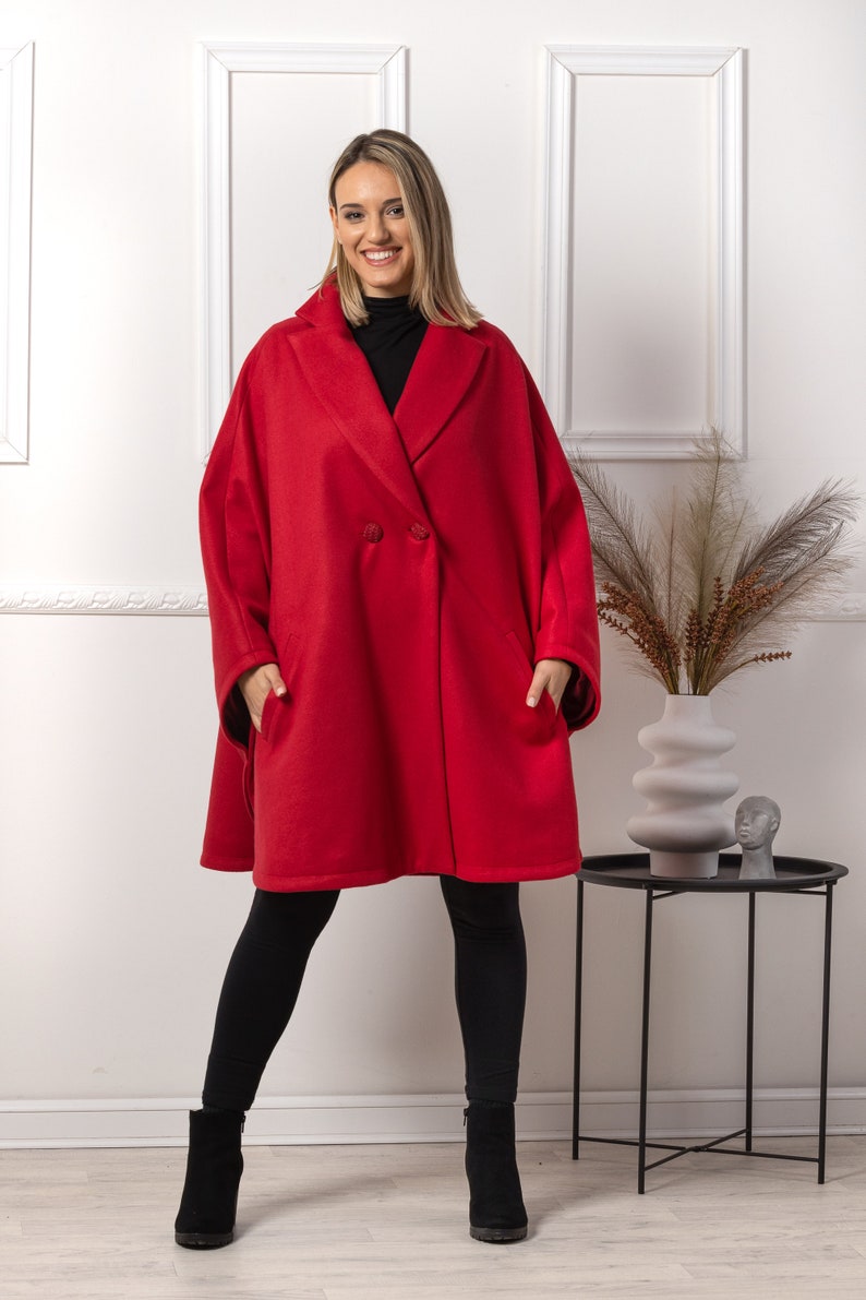 May include: A double-breasted red coat with large lapels and pockets. The coat is knee-length and worn with black leggings and ankle boots. The background is a white wall with decorative trim.