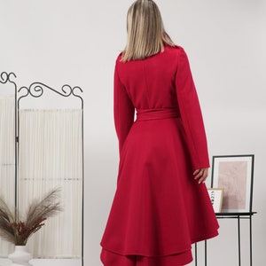 May include: A vibrant red coat featuring a high-low hemline with layered fabric panels, long sleeves, and a matching tie belt at the waist. The coat is paired with black ankle boots, showcasing a structured silhouette and a smooth, solid finish.