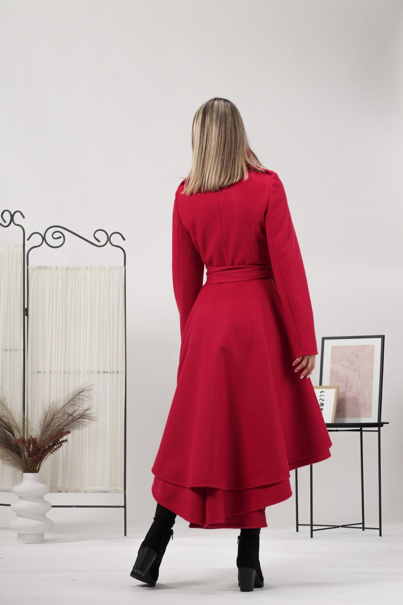 May include: A vibrant red coat featuring a high-low hemline with layered fabric panels, long sleeves, and a matching tie belt at the waist. The coat is paired with black ankle boots, showcasing a structured silhouette and a smooth, solid finish.