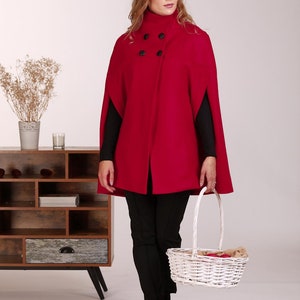 May include: A vibrant red cape featuring a high collar and four black buttons arranged in a double-breasted style. The garment has arm slits on the sides and is worn over a black long-sleeved top and black trousers, paired with a white woven basket.