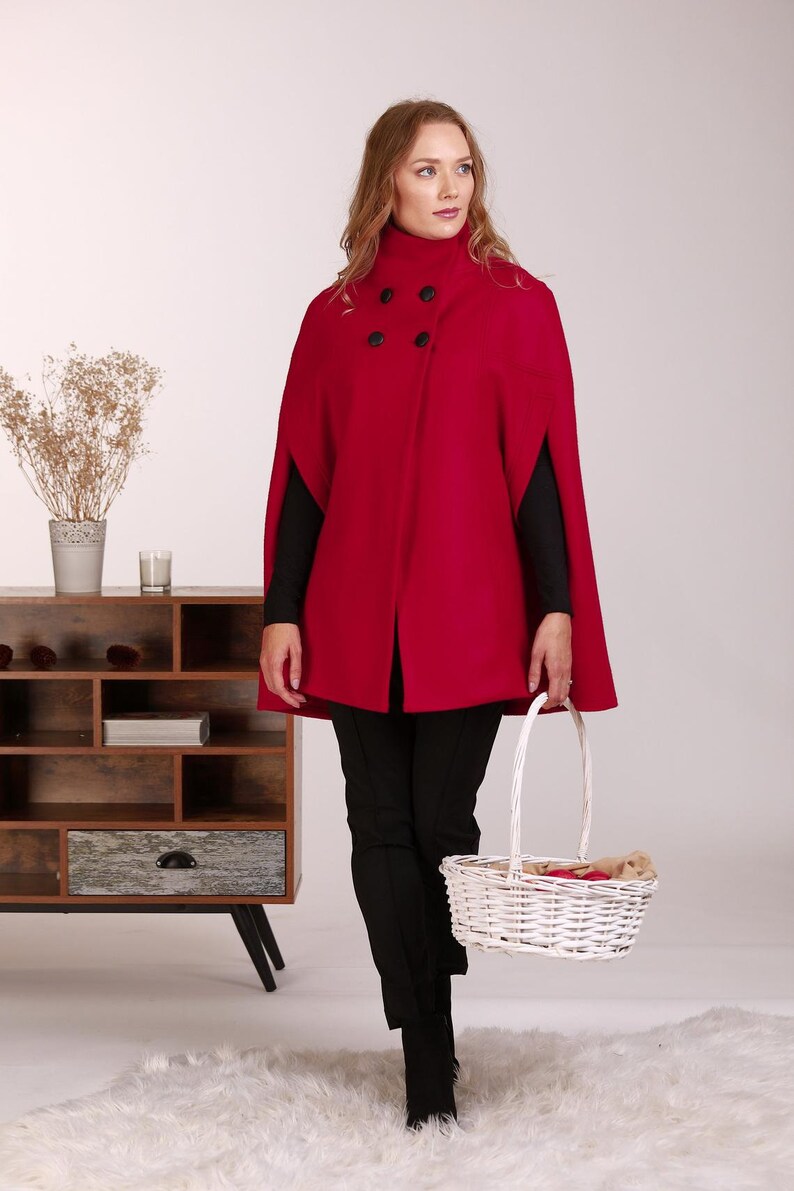 May include: A vibrant red cape featuring a high collar and four black buttons arranged in a double-breasted style. The garment has arm slits on the sides and is worn over a black long-sleeved top and black trousers, paired with a white woven basket.