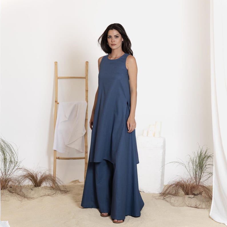 May include: A sleeveless, floor-length, denim blue dress with a layered skirt. The dress has a simple, round neckline and a relaxed fit. The model is standing in a studio setting with a bamboo ladder and a white cloth.