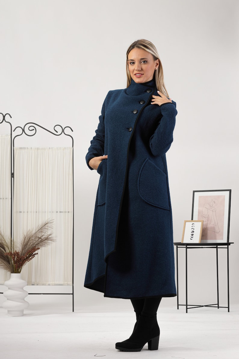 May include: A long, navy blue coat with a high collar and large, round buttons. The coat features two front pockets and a black trim along the edges. The coat is worn with black pants and boots.