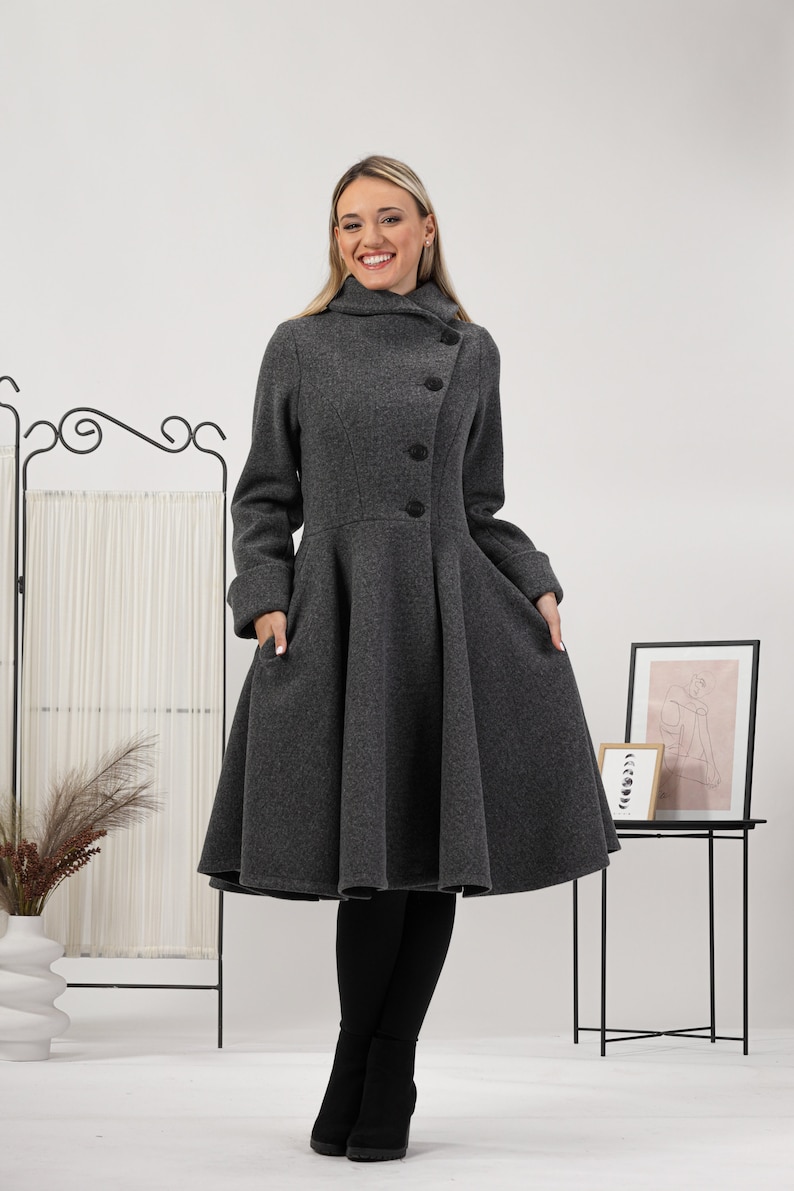 May include: A charcoal grey coat featuring a high collar, long sleeves with folded cuffs, and a flared skirt silhouette. The coat has a diagonal button closure with four black buttons and two side pockets, extending to just below the knee.