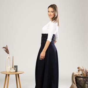 May include: A woman in a navy blue, high-waisted skirt with button detailing and a white long-sleeved top. The skirt is floor-length and flared. A small wooden table with a vase and candles is to the left. The skirt's length is approximately 100 cm.
