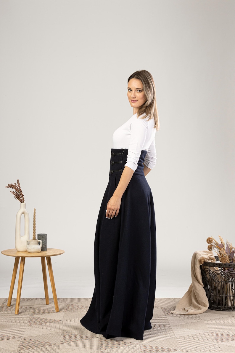 May include: A woman in a navy blue, high-waisted skirt with button detailing and a white long-sleeved top. The skirt is floor-length and flared. A small wooden table with a vase and candles is to the left. The skirt's length is approximately 100 cm.