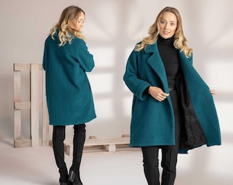 May include: A teal-colored coat with a classic lapel collar and long sleeves. The coat is knee-length and has a relaxed fit. The model is wearing black pants and black boots. The coat's interior lining is black.