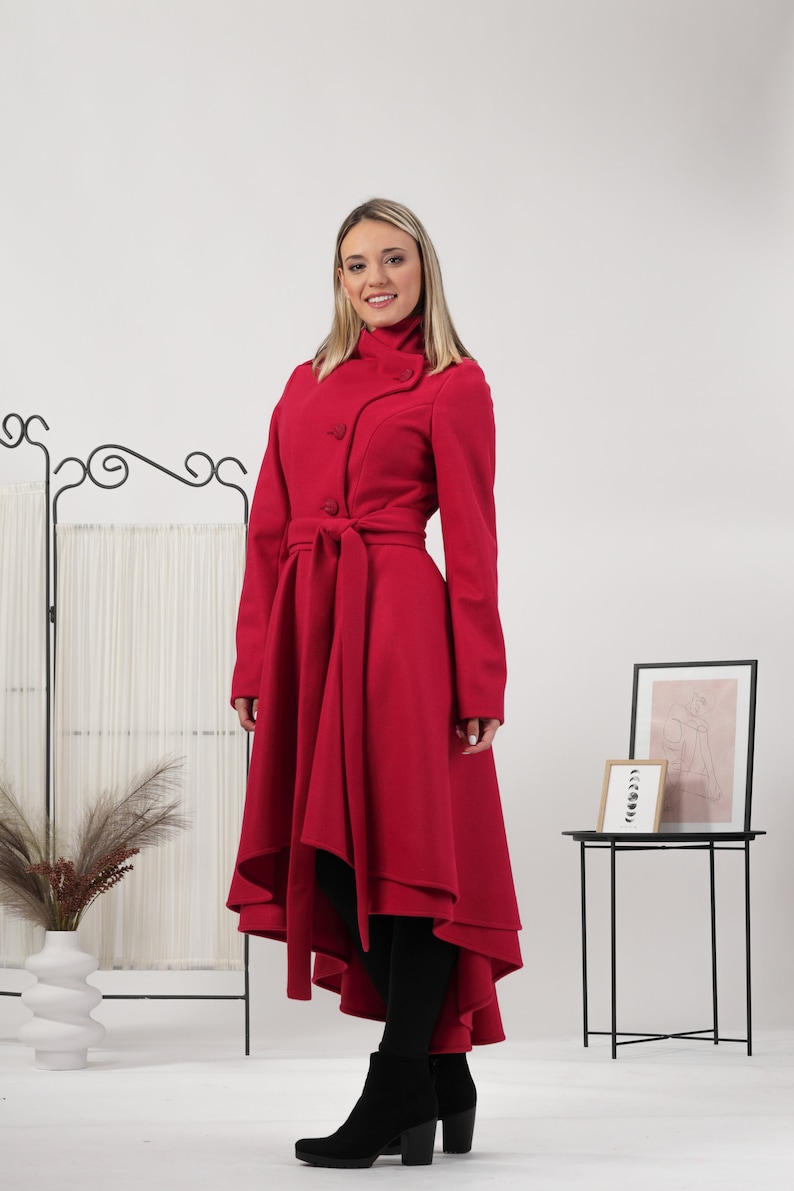 May include: A vibrant red long-sleeved coat featuring a high collar, a button-front closure, and a matching fabric tie belt at the waist. The coat has a high-low hemline that is shorter in the front and longer in the back, creating a flared silhouette.