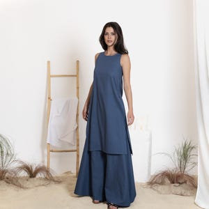 May include: A sleeveless, blue dress with a layered design. The dress has a long, flowing skirt and a high neckline. The model is standing in front of a white background with a wooden ladder and some plants.