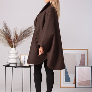 May include: A brown cape coat with a wide collar and a flowing silhouette. The coat has a simple design with a slightly rounded hemline. The model is wearing black leggings and black heeled boots. The background includes a small black side table and framed artwork.