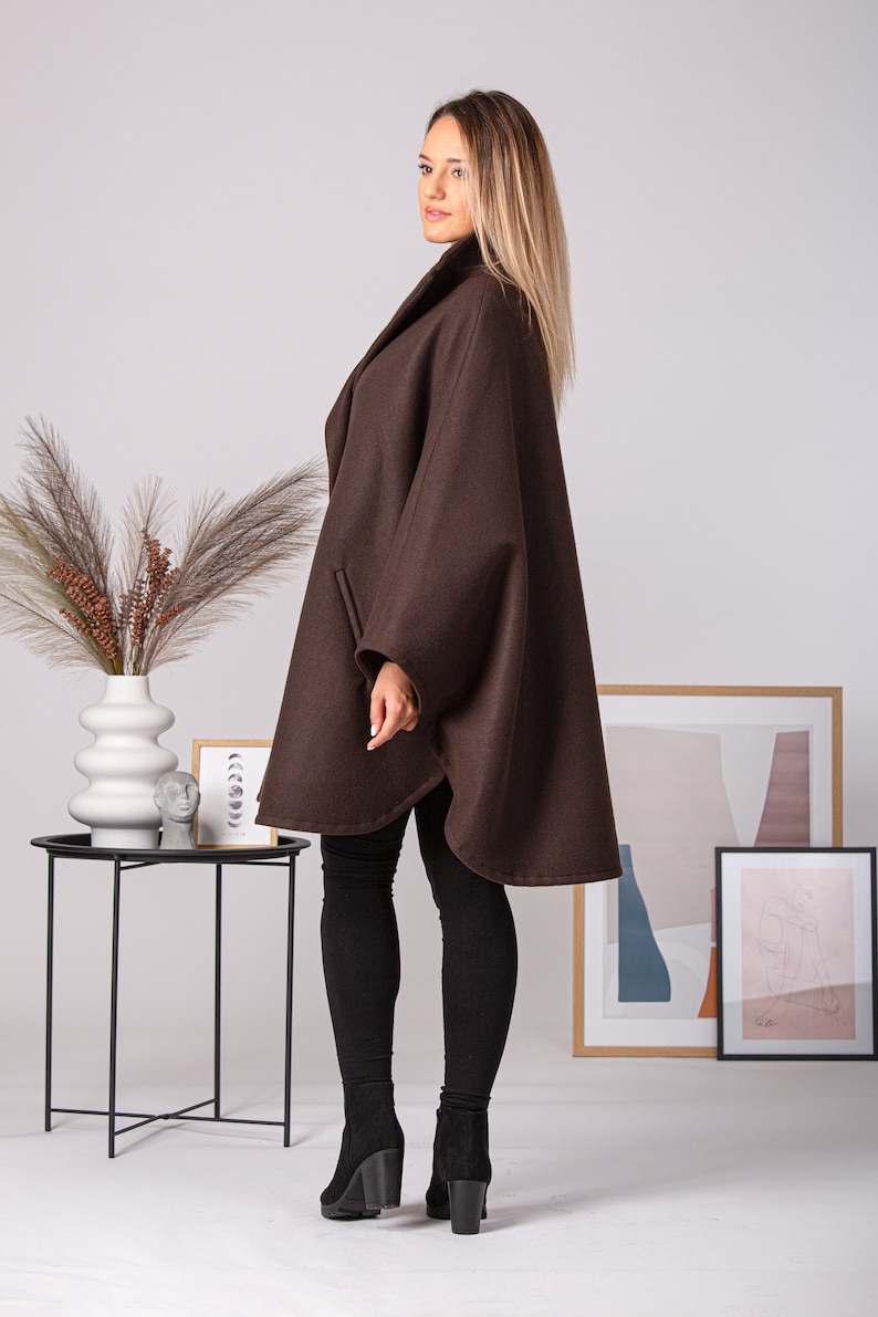 May include: A brown cape coat with a wide collar and a flowing silhouette. The coat has a simple design with a slightly rounded hemline. The model is wearing black leggings and black heeled boots. The background includes a small black side table and framed artwork.