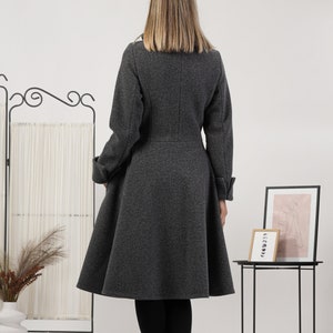 May include: A charcoal grey knee-length coat with a fitted bodice and a flared skirt. The coat features long sleeves with folded cuffs, a high collar, and a vertical seam running down the centre of the back. The fabric has a textured, wool-like finish.