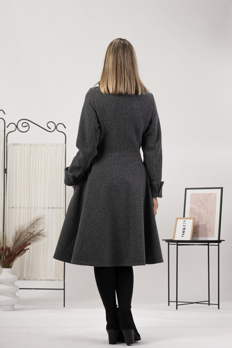May include: A charcoal grey knee-length coat with a fitted bodice and a flared skirt. The coat features long sleeves with folded cuffs, a high collar, and a vertical seam running down the centre of the back. The fabric has a textured, wool-like finish.