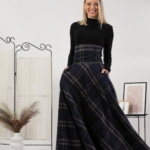 May include: A woman in a black top and a long, plaid skirt. The skirt has a high waist with buttons and pockets. The plaid pattern is in blue, grey, and brown. The woman is smiling. The background is white. The skirt's length is approximately 100 cm.