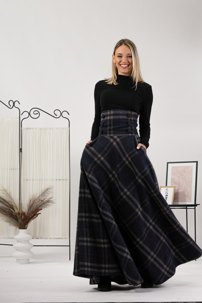 May include: A woman in a black top and a long, plaid skirt. The skirt has a high waist with buttons and pockets. The plaid pattern is in blue, grey, and brown. The woman is smiling. The background is white. The skirt's length is approximately 100 cm.