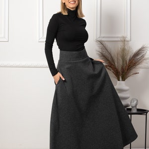 May include: A woman wearing a black turtleneck long-sleeve shirt and a dark gray maxi skirt with pockets. The skirt is a solid color and has a high waist. The woman is smiling and standing in front of a white wall.