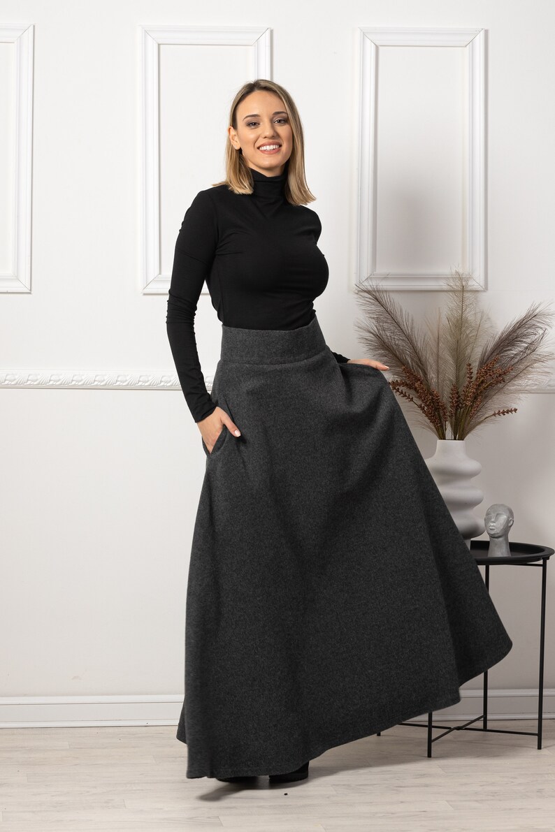 May include: A woman wearing a black turtleneck long-sleeve shirt and a dark gray maxi skirt with pockets. The skirt is a solid color and has a high waist. The woman is smiling and standing in front of a white wall.
