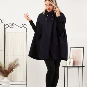 May include: A dark navy blue wool-blend cape featuring a high collar and four large, round black buttons arranged in a double-breasted style. The garment has wide arm openings and a structured, flared silhouette that falls to the mid-thigh.