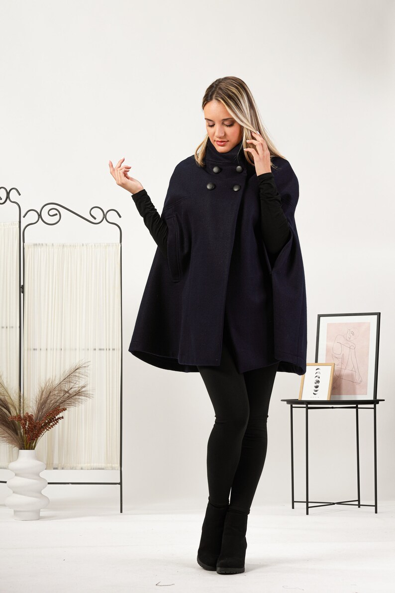 May include: A dark navy blue wool-blend cape featuring a high collar and four large, round black buttons arranged in a double-breasted style. The garment has wide arm openings and a structured, flared silhouette that falls to the mid-thigh.