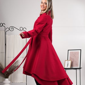 May include: A vibrant red coat featuring a high-low hemline, long sleeves, and a matching fabric tie belt at the waist. The garment is worn with black leggings and black heeled boots, showcasing a flared silhouette that extends to the mid-calf.