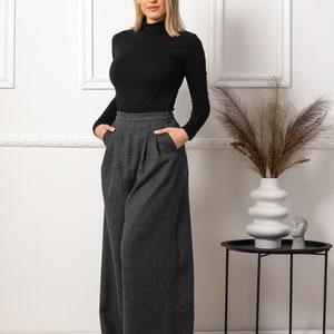 May include: A woman wearing a black turtleneck top and wide-leg, gray wool trousers. The pants have a high waist and pleats. The outfit is simple and elegant, suitable for various occasions. The background is a white wall with decorative frames.
