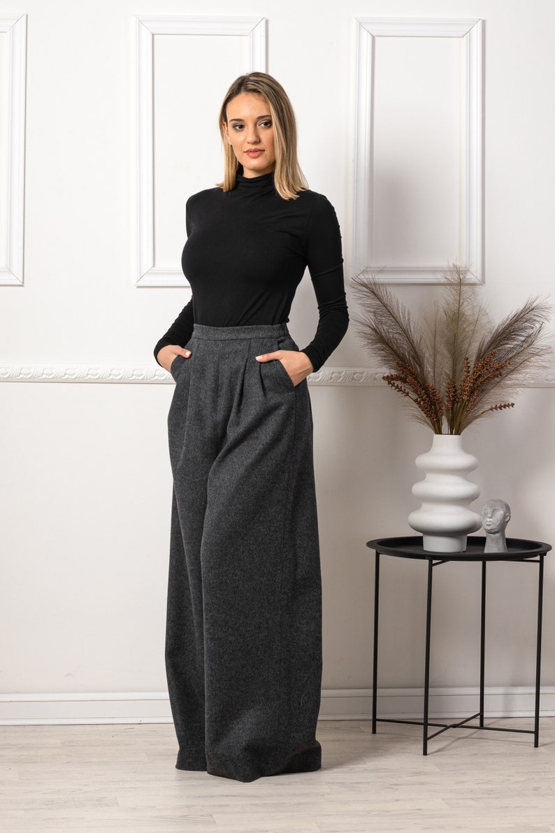 May include: A woman wearing a black turtleneck top and wide-leg, gray wool trousers. The pants have a high waist and pleats. The outfit is simple and elegant, suitable for various occasions. The background is a white wall with decorative frames.