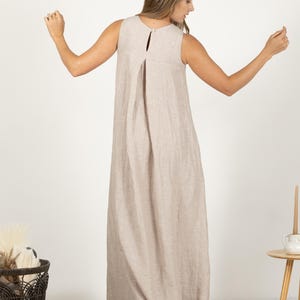 K&ouml;nnte beinhalten: Ein &auml;rmelloses, bodenlanges Kleid in einem neutralen Beigeton. Das Kleid hat ein Schl&uuml;ssellochdetail am R&uuml;cken und eine plissierte Vorderseite. Das Model tr&auml;gt das Kleid mit ausgestreckten Armen. Das Kleid scheint aus Leinen zu sein.