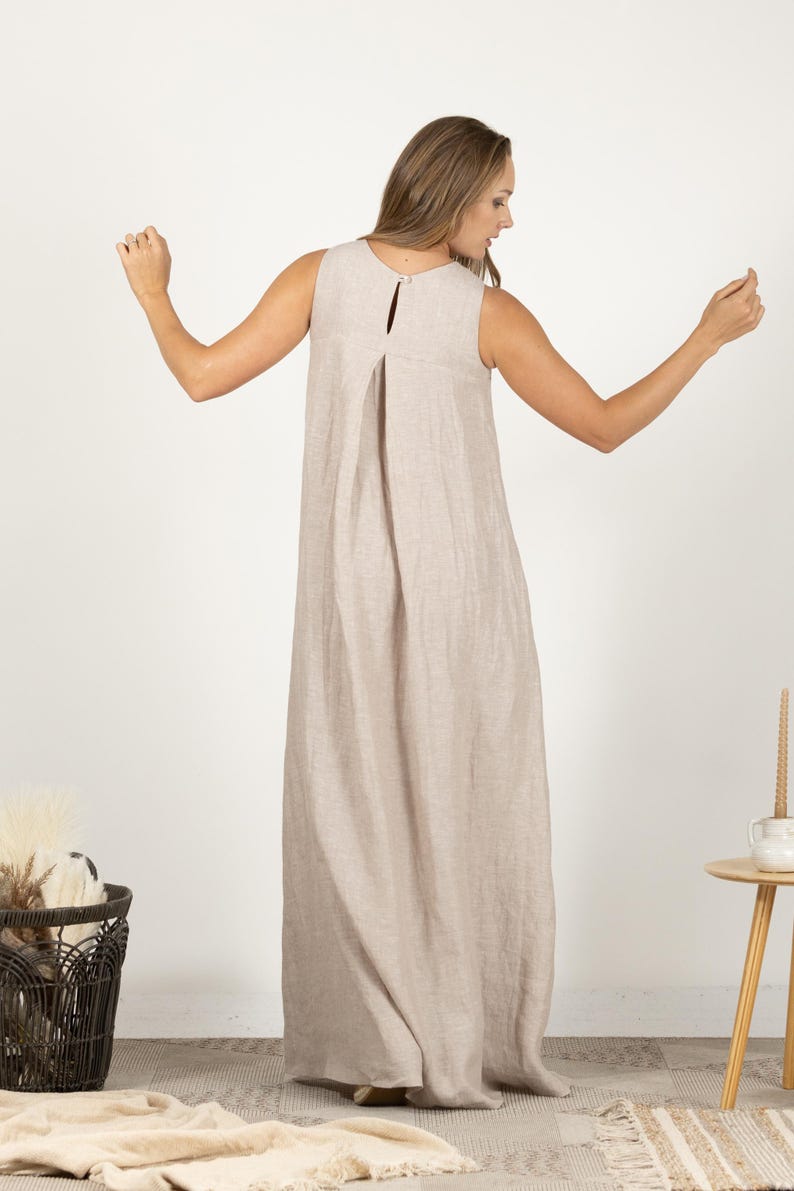 K&ouml;nnte beinhalten: Ein &auml;rmelloses, bodenlanges Kleid in einem neutralen Beigeton. Das Kleid hat ein Schl&uuml;ssellochdetail am R&uuml;cken und eine plissierte Vorderseite. Das Model tr&auml;gt das Kleid mit ausgestreckten Armen. Das Kleid scheint aus Leinen zu sein.