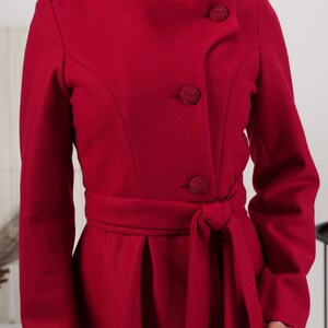 May include: A vibrant red coat featuring a high collar, long sleeves, and a belted waist. The front closure includes three large, textured, circular buttons. The coat has structured shoulders and a tailored fit with a tie-waist belt.