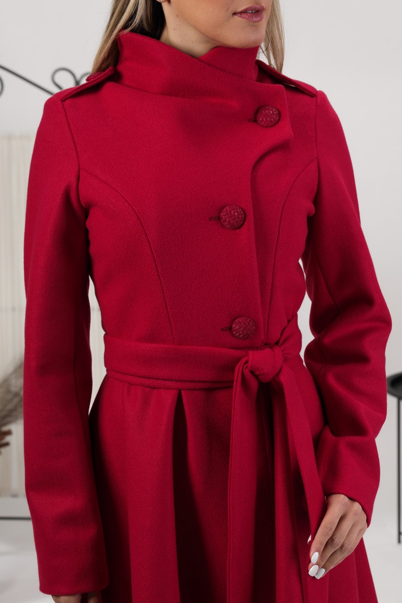 May include: A vibrant red coat featuring a high collar, long sleeves, and a belted waist. The front closure includes three large, textured, circular buttons. The coat has structured shoulders and a tailored fit with a tie-waist belt.