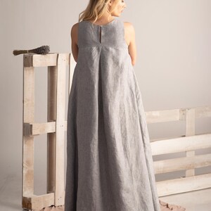 K&ouml;nnte beinhalten: Ein bodenlanges, &auml;rmelloses graues Leinenkleid mit Schl&uuml;sselloch-R&uuml;cken. Das Kleid hat ein schlichtes, elegantes Design mit einer leichten A-Linien-Silhouette. Ein B&uuml;ndel getrockneter Lavendel liegt auf einer Holzpalette im Hintergrund.