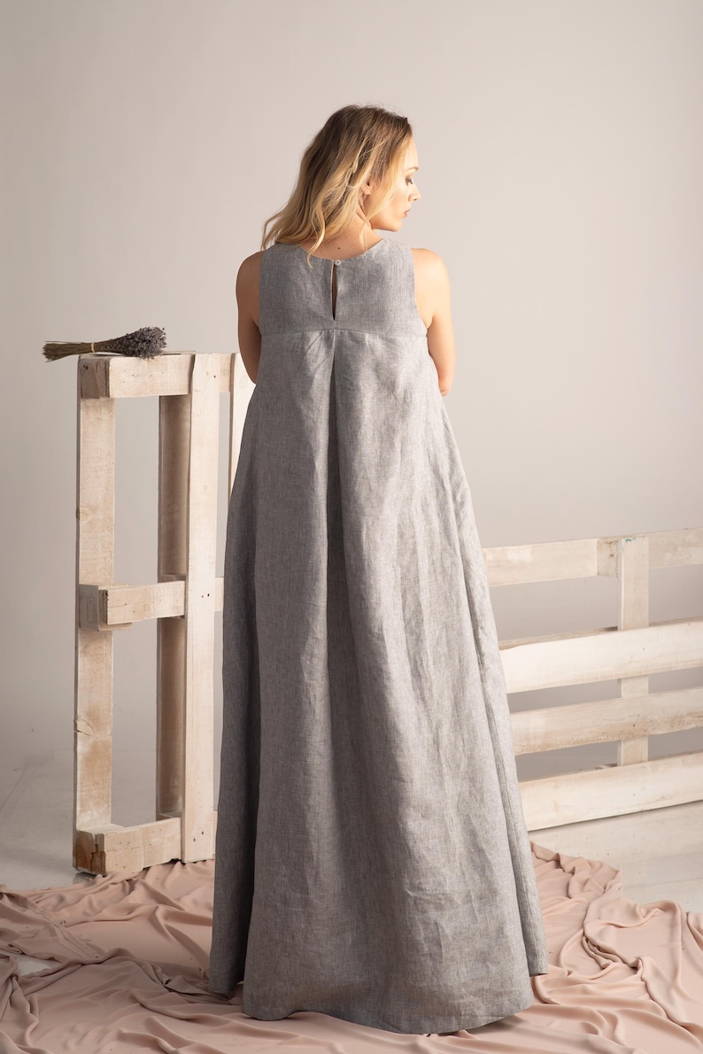 K&ouml;nnte beinhalten: Ein bodenlanges, &auml;rmelloses graues Leinenkleid mit Schl&uuml;sselloch-R&uuml;cken. Das Kleid hat ein schlichtes, elegantes Design mit einer leichten A-Linien-Silhouette. Ein B&uuml;ndel getrockneter Lavendel liegt auf einer Holzpalette im Hintergrund.