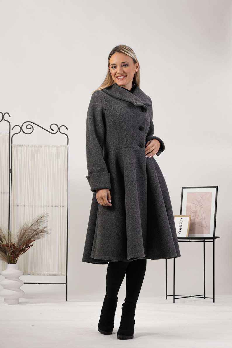 May include: A charcoal grey wool-blend coat featuring a wide folded collar, three large black buttons down the front, long sleeves with cuffs, and a flared, knee-length skirt silhouette. The coat is worn over black leggings and black ankle boots.