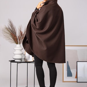 May include: A brown cape with a high collar. The cape is made of a soft material and is worn over black leggings and black boots. The model is standing in a well-lit room.