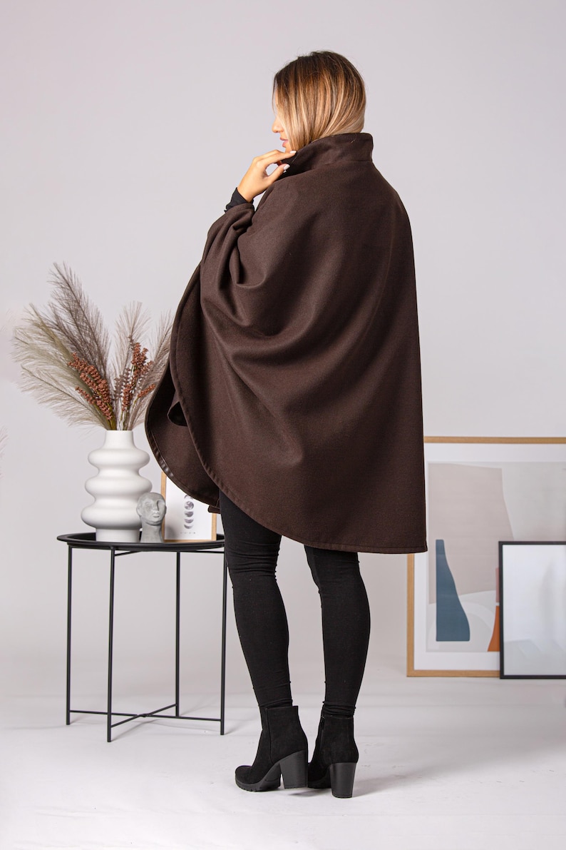 May include: A brown cape with a high collar. The cape is made of a soft material and is worn over black leggings and black boots. The model is standing in a well-lit room.