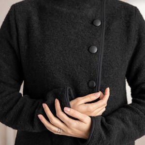May include: A black wool coat with a high collar and three large, round buttons. The coat has a textured surface and black trim along the edges and cuffs. The sleeves are long, and the coat appears to be a warm, stylish outerwear garment.