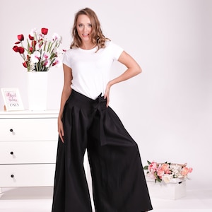 May include: A person wearing a white t-shirt and wide-leg black trousers with a matching tie. The trousers are high-waisted and flowy. The background includes a white chest of drawers, flowers, and a sign that says "Good vibes only."