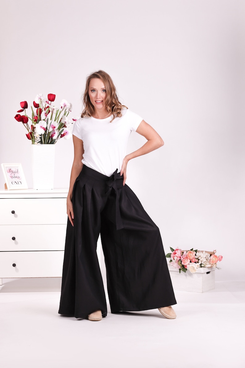 May include: A person wearing a white t-shirt and wide-leg black trousers with a matching tie. The trousers are high-waisted and flowy. The background includes a white chest of drawers, flowers, and a sign that says "Good vibes only."