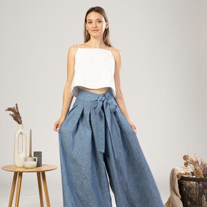 May include: A woman in a white crop top and wide-leg, high-waisted blue trousers with a matching tie. The trousers are made of a light fabric. The outfit is casual and stylish.