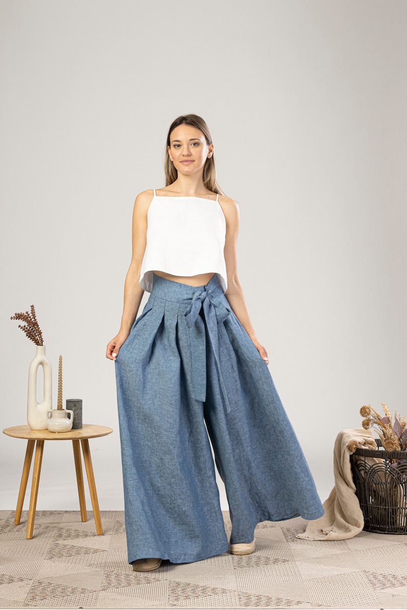 May include: A woman in a white crop top and wide-leg, high-waisted blue trousers with a matching tie. The trousers are made of a light fabric. The outfit is casual and stylish.