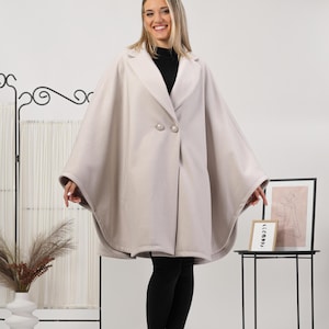 May include: A light beige cape coat with a double-breasted closure and two large buttons. The coat has a wide, flowing design and is worn over a black top and leggings. The model is smiling.